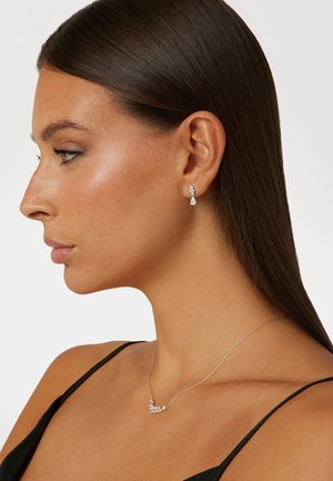 Collier en argent avec des pierres transparentes dans un design courbé, associé à des boucles d'oreilles pendantes ornées de gouttes et d'accents en pierre.
