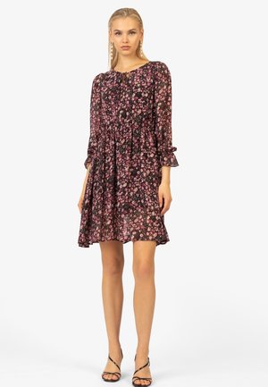 Robe fleurie noire avec des motifs roses et blancs, manches longues avec des poignets froncés, taille cintrée et jupe évasée, assortie à des sandales noires à lanières.