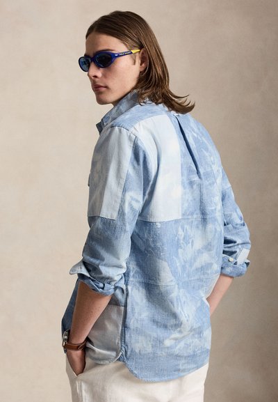 Persoană tânără cu păr până la umeri poartă ochelari de soare cu lentile albastre, o cămașă albastru deschis cu model și pantaloni crem, cu mâinile în buzunare.