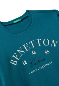 Tyrkysový svetr vyrobený z jemné látky má žebrovaný kulatý výstřih a tištěné logo "BENETTON" s rokem a detaily značky v kontrastních barvách.