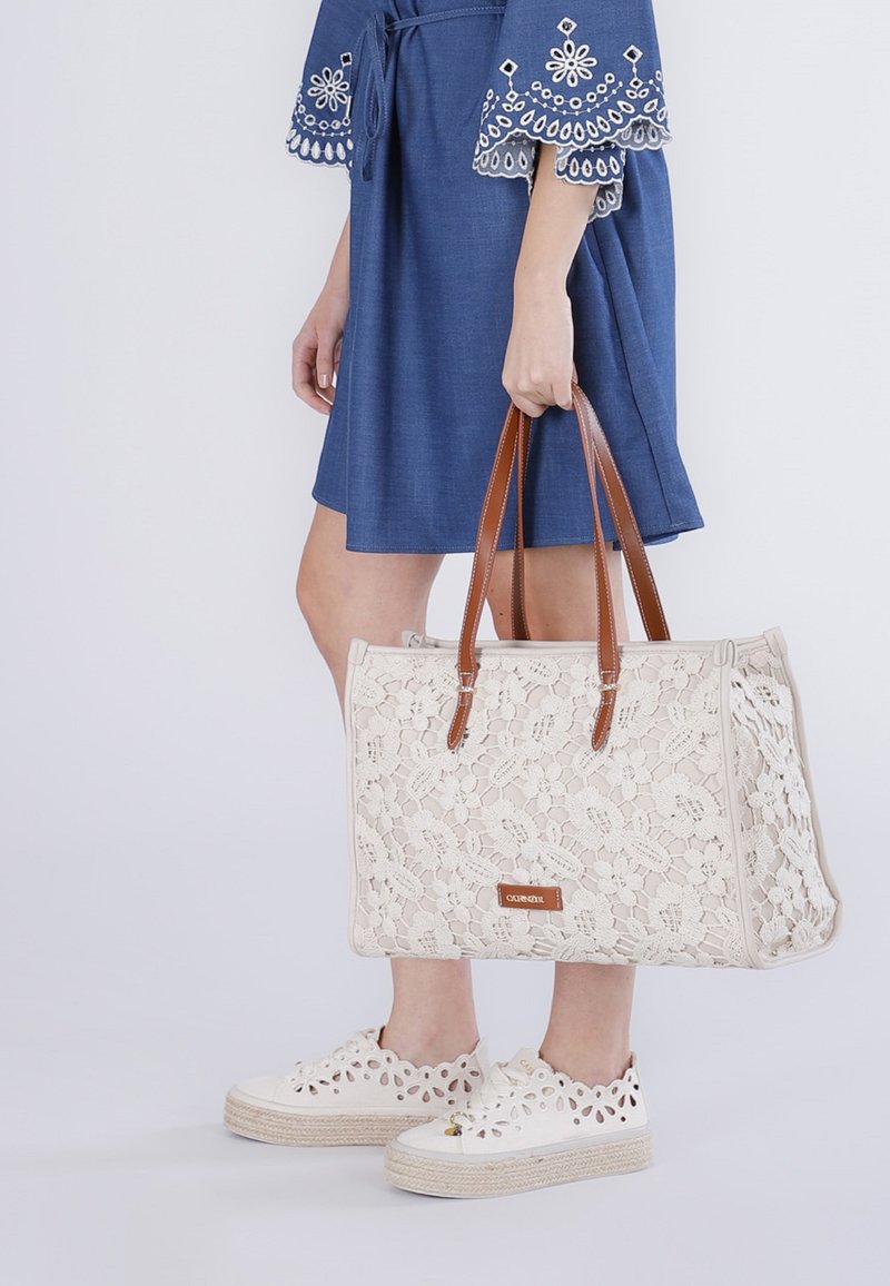 Kvinde i blå kjole med broderede ærmer holder en cremefarvet blondertaske med brune håndtag, iført hvide platformssneaks med udskæringer.