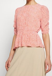 Červená a bílá květinová blůza s peplum lemem, krátkými nafouklými rukávy a kulatým výstřihem. Lehká tkanina s jemnou strukturou.