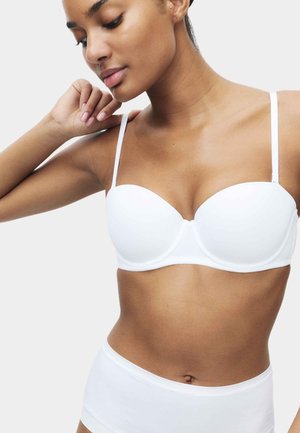 Mujer con sujetador blanco acolchado y ropa interior de talle alto a juego, posando con un brazo levantado y mirando hacia abajo.