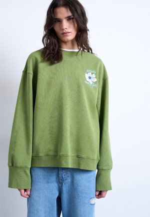 Jonge vrouw met donker haar tot op de schouders draagt een oversized groene trui met wit bloemmotief en lichtblauwe gescheurde spijkerbroek tegen een witte achtergrond.