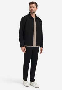 Zwarte jas met een kraag, beige shirt eronder, zwarte broek en witte sneakers. De jas heeft een ritssluiting en zakken.