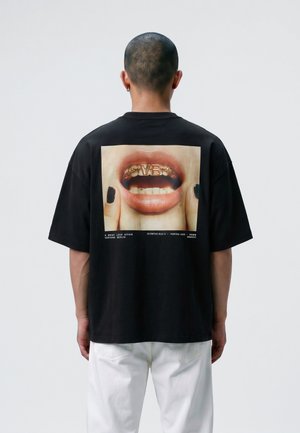 Osoba z krótkimi włosami, ubrana w czarny, oversize t-shirt z dużą grafiką ust i złotych zębów na plecach oraz białe spodnie.