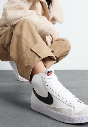 Person iført beige bukser og en cremefarvet fleecejakke, der sidder på hug for at vise hvide high-top Nike sneakers med en sort swoosh.