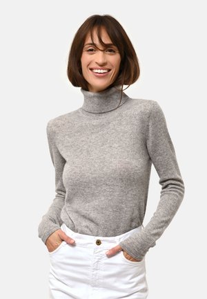 Maglione grigio a collo alto in tessuto morbido con maniche lunghe, caratterizzato da una silhouette aderente. Il modello indossa pantaloni bianchi, con le mani nelle tasche.