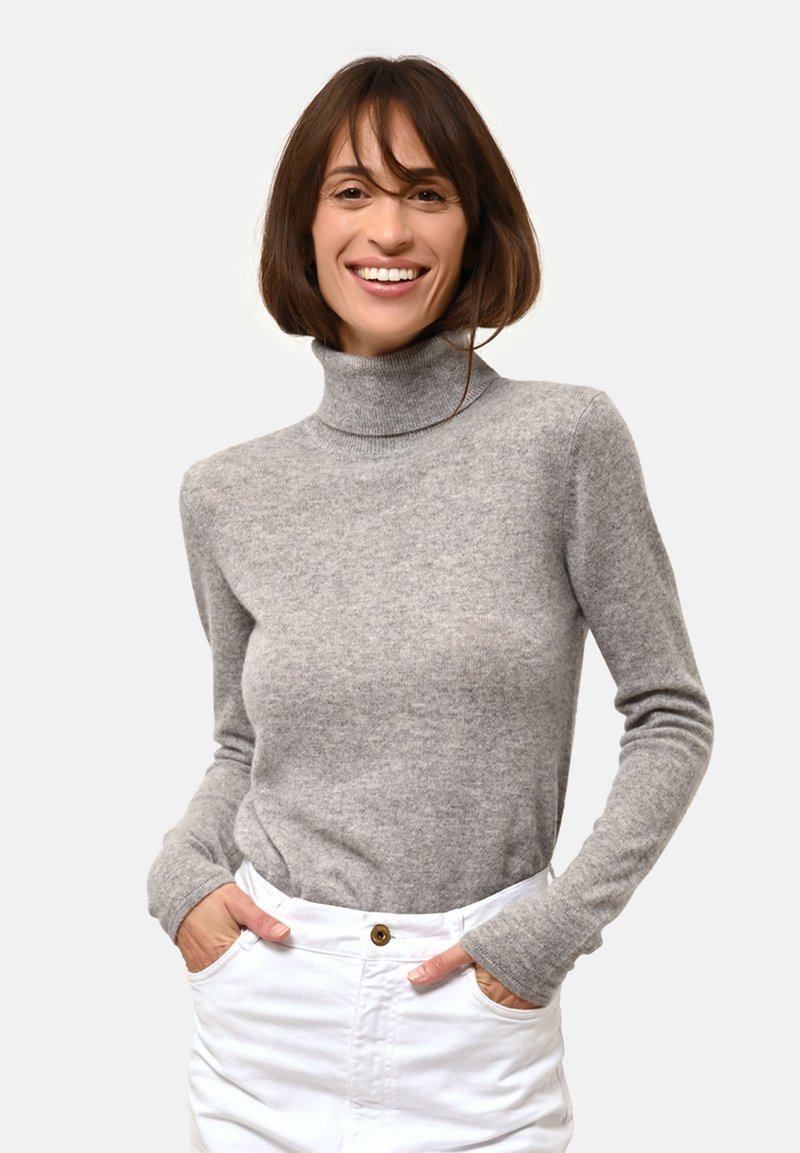 Šedý rolák z měkké tkaniny s dlouhými rukávy a přiléhavým střihem. Model má na sobě bílé kalhoty a ruce v kapsách.