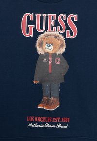 Felpa navy con una grafica di un orso che indossa un cappotto scuro, un maglione rosso, jeans e stivali marroni, con "GUESS" e dettagli del marchio sottostanti.