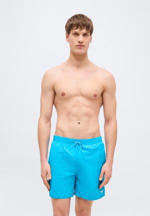 Mladý muž stojící čelem vpřed, bez trička, oblečený ve světlých modrých plavkách se stahovací šňůrkou v pase.