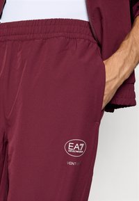 Pantaloni atletici bordo cu un tiv elastic, buzunare laterale și un logo alb care recită "EA7 Emporio Armani VENTUS." Textură netedă.