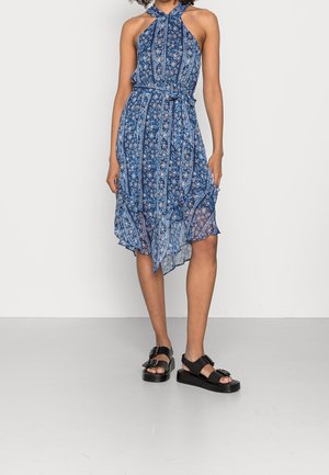 Vrouw draagt een blauwe gebloemde mouwloze halterjurk met een asymmetrische zoom en zwarte sandalen met gesp tegen een witte achtergrond.