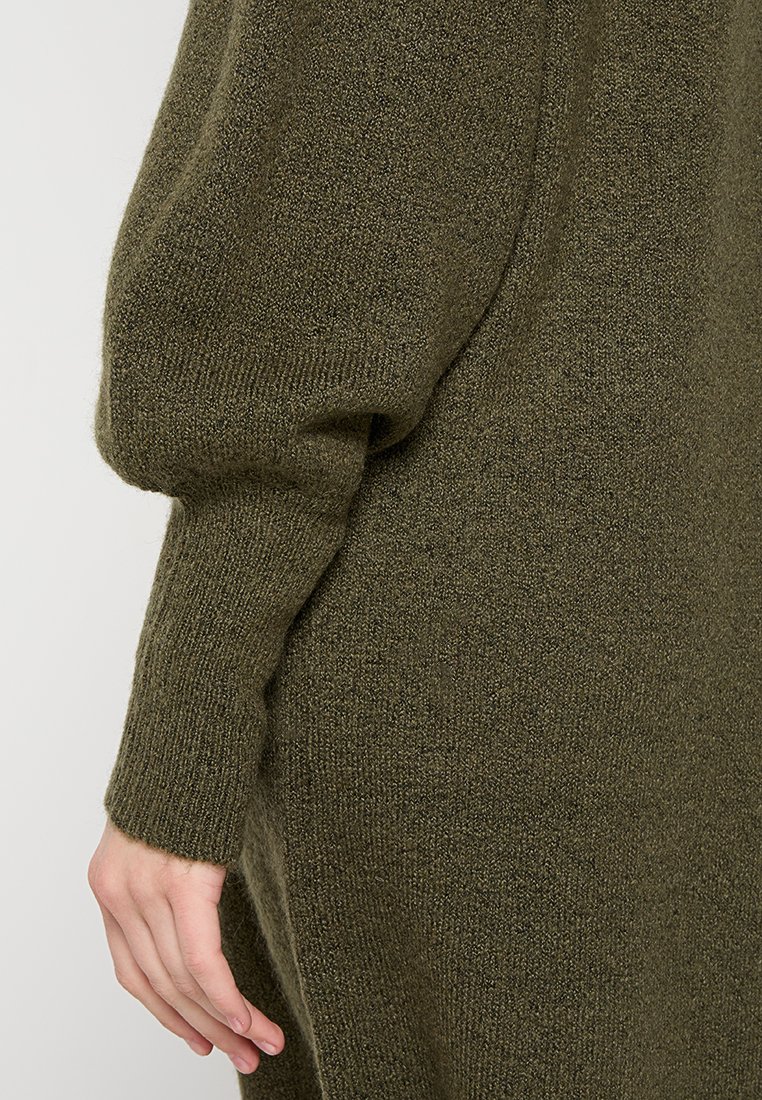Dunkelgrüner Strickpullover mit lockerer Passform, gerippten Bündchen und strukturiertem Stoff. Sichtbare Hand, die an der Seite ruht.