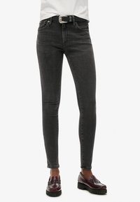 Jeans skinny noirs en denim, avec une taille mi-haute, cinq poches et une texture légèrement délavée, complétés par une ceinture à boucle argentée.