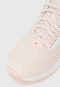 Zapatilla deportiva de color rosa claro con parte superior de malla texturizada, cordones en contraste y suela blanca. Cuenta con un logotipo en el lateral y sutiles detalles de diseño.