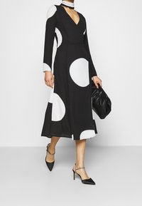 Schwarzes Kleid mit langen Ärmeln, V-Ausschnitt und großen weißen Punkten. Getragen mit schwarzen Stiletto-Absätzen und einer schwarzen Tasche. Glatte Stofftextur.