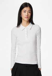 Bílá polokošile s dlouhým rukávem v žebrované struktuře, vybavená límečkem a pětice knoflíků na légu. Natažená k černých kalhotám s vysokým pasem.