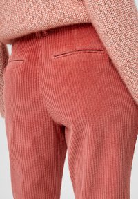 Puuvillapantonit pölyisen vaaleanpunaisina, joissa on pystyribbitekstuuri, kaksi takataskua ja istuva muotoilu. Neule osittain näkyvissä yllä.