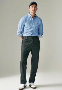 Camisa de botones azul claro, pantalones oscuros grises y zapatillas deportivas blancas. La tela parece suave con una textura sutil; se lleva una gorra de béisbol.