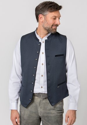 Mann trägt ein weißes Hemd mit Knöpfen, eine dunkelblaue gemusterte Weste und graue Wildlederhosen mit dekorativer Gürtelschnalle, schaut seitlich und lächelt.