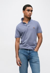 Krátky rukáv, polokošeľa s golierom v svetlofialovej bavlne, zdobená malým vyšívaným motívom na hrudi. Nosí sa s modrými džínsami z denimu.