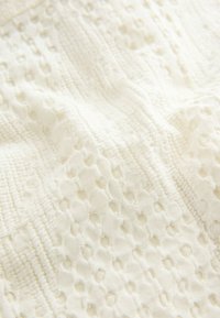 Prim-plan al unei țesături moi, de culoare crem, cu modele complicate din dantelă și desene țesute, texturate.