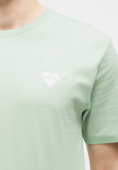 Μεταξωτό βαμβακερό πράσινο ανοιχτό μπλουζάκι με έμβλημα καρδιάς και βέλους και κείμενο "THE KOOPLES" στο αριστερό στήθος, φορεμένο από ένα άτομο.