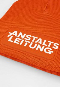 Orange knit beanie featuring a textured fabric and a prominent white patch with the text "ANSTALTSLEITUNG" embroidered on it.