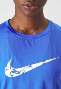Acercamiento al pecho de una persona que lleva una camisa azul con un logotipo de 'swoosh' blanco estilizado de Nike.