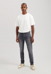 Tricou alb cu mânecă scurtă, combinat cu blugi negri slim-fit și adidași crem cu accente galbene. Modelul stă în fața unui fundal neutru.
