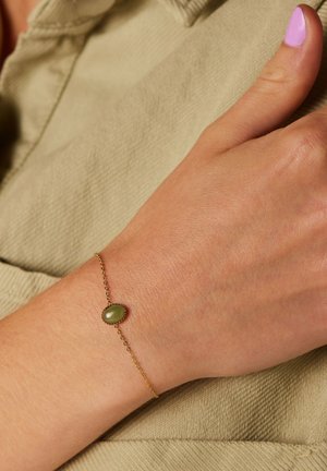 Bracelet en chaîne dorée avec un centre en pierre verte ovale, porté sur une main contre un fond en tissu beige texturé.