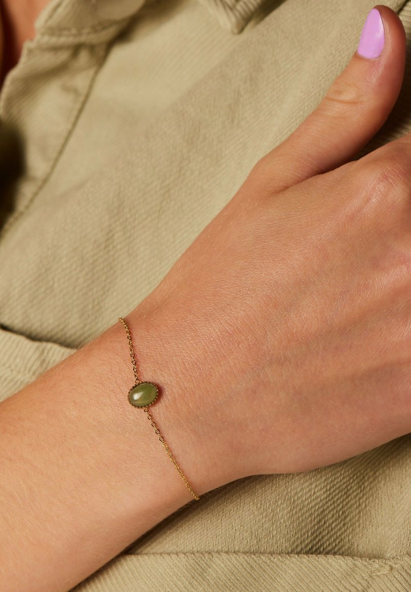 Gouden kettingarmband met een ovale groene steen als middelpunt, gedragen aan een hand tegen een textuur van beige stof als achtergrond.