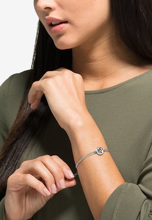 Silberne Armbanduhr mit einem geflochtenen Kettendesign, das einen runden Metalleinsatz aufweist. Die Hand der Person ist teilweise sichtbar und zeigt die Passform des Produkts.