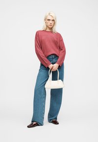 Jeune femme aux cheveux blond platine portant un pull rouge ample, un jean pattes d'éléphant bleu, des mocassins marron, et tenant un petit sac à main blanc.