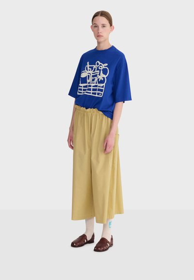 Femme portant un t-shirt graphique bleu oversize, un pantalon beige large et raccourci, des chaussettes blanches et des sandales en cuir marron, debout devant un fond uni.