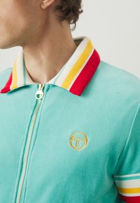 Veste zippée en polaire aqua avec col côtelé, ornée de rayures rouges, jaunes et bleues ; comprend un logo brodé sur la poitrine.
