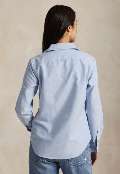 Kvinde med langt mørkt hår iført en lyseblå langærmet skjorte og blå jeans, stående med ryggen til mod en ensfarvet beige baggrund.