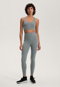 Svetlošedý športový podprsenka a zodpovedajúce legíny s vysokým pásom vyrobené z elastického materiálu; skombinované s čiernymi teniskami; hladká textúra, minimalistický dizajn.