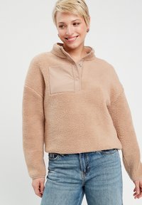 Pullover en polaire beige clair avec un col zippé à mi-hauteur, présentant un tissu texturé et une poche poitrine en matériau lisse.