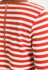 Czerwono-biała tkanina w paski o miękkiej fakturze, z widocznym złotym zamkiem z tyłu i okrągłym dekoltem.