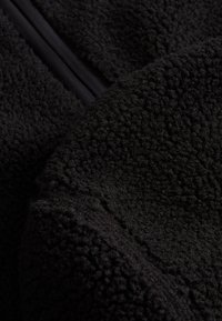 Zwart fleece textiel met een gestructureerd oppervlak en een zachte, pluizige uitstraling. Bevat een gladde ritsdetail langs één rand.