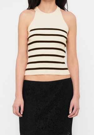 Vrouw die een mouwloze beige top met horizontale zwarte strepen draagt, gecombineerd met een zwarte kanten rok, geknipt ter hoogte van de taille tegen een effen achtergrond.