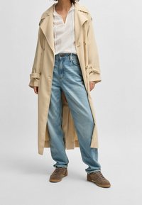 Osoba w beżowym długim płaszczu, białym teksturowanym hoodie, luźnych jasnoniebieskich dżinsach i brązowych zamszowych sneakersach, stojąca na szarym podłodze.