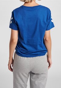 Camiseta de algodón azul con mangas cortas, con acentos en chevrón blancos en los hombros. Combinada con pantalones de chándal grises, mostrando un ajuste casual.