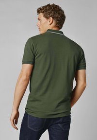 Olivgrön polo med en figurnära design, korta ärmar och smala vita ränder på kragen. Bärs tillsammans med mörkblå jeans.