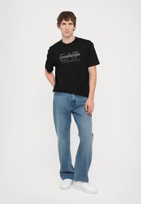Black cotton t-shirt with E.A. Emporio Armani logo in gray, paired with blue jeans and white sneakers, presented in a neutral background.