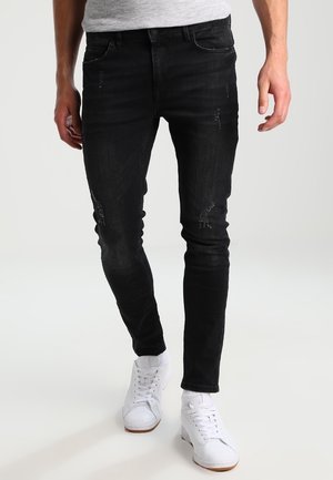 Homme portant un jean noir slim déchiré et des baskets blanches, debout devant un fond clair uni.