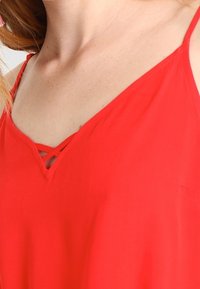 Haut rouge sans manches avec un col en V et des détails croisés au niveau de l'encolure ; tissu lisse et coupe décontractée.