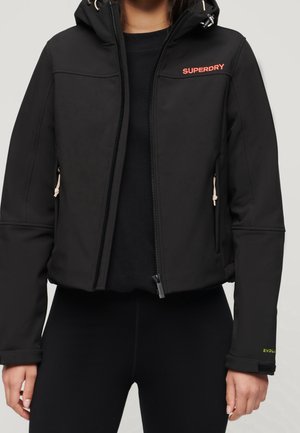 Femme portant une veste noire zippée à capuche, logo rose "SUPERDRY" sur la poitrine, tirettes de fermeture éclair beiges et leggings noirs.